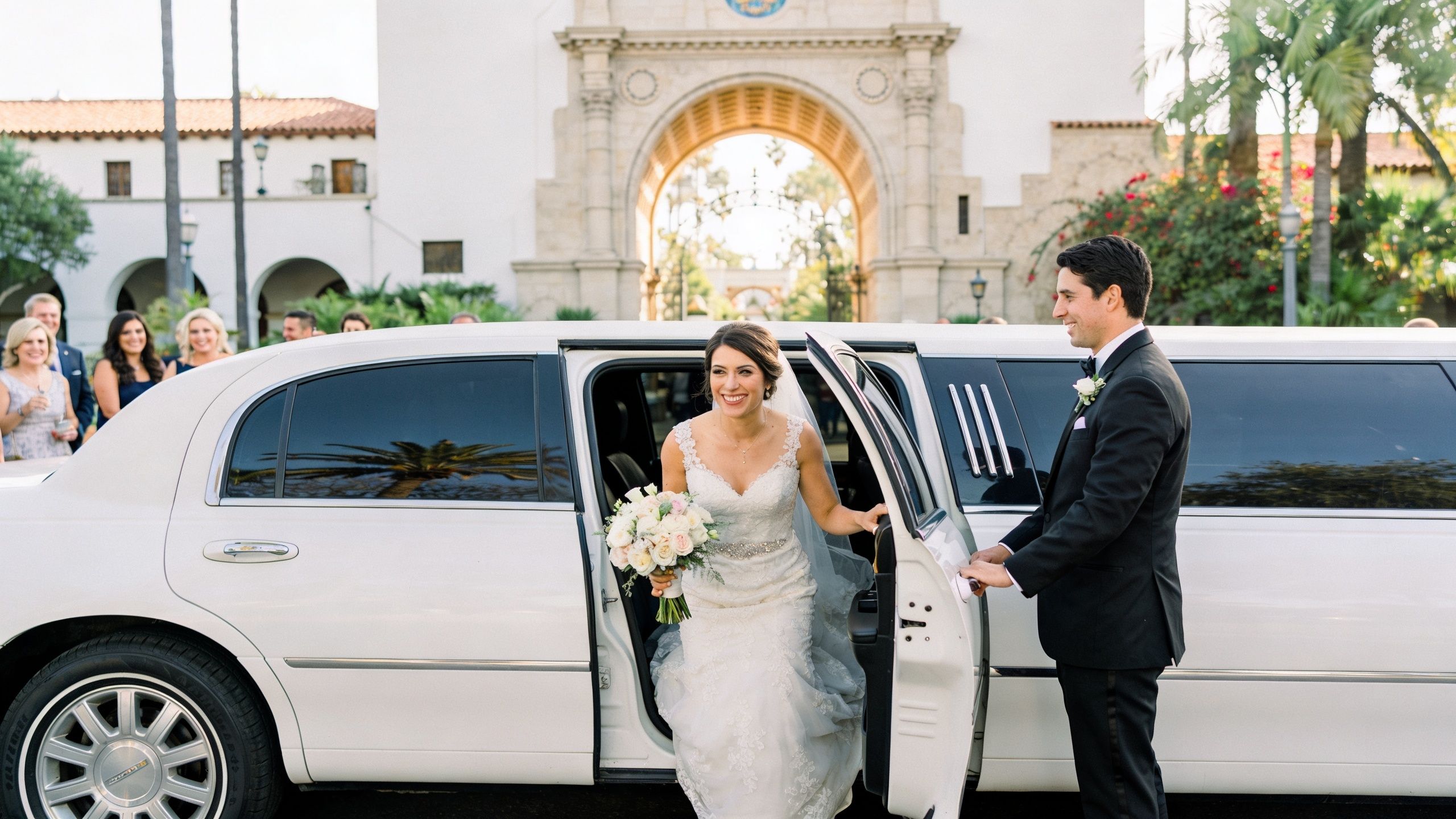 A smiling bride in a white wedding gown steps out of a limousine while her groom watches.